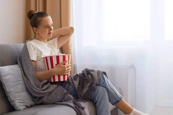 Horizontal shot of sad woman wearing white t shirts wrapped in blanket watching movie and touching neck, sitting on sofa, expressing sadness and sorrow.