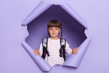 Örgü örgülü, elinde sırt çantasıyla okula gitmeye hazır bir şekilde kameraya bakan çekici küçük bir kızın kapalı çekimleri..