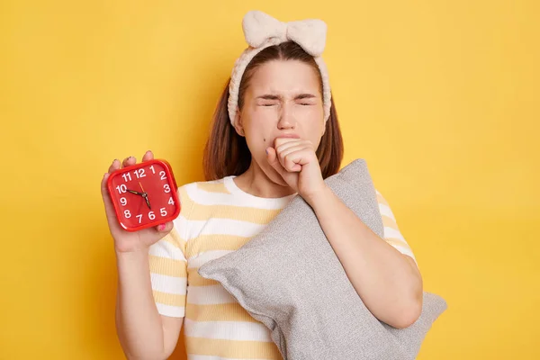 Indoor shot of sleepy young female with dark hair holding alarm clock and embracing pillow with closed eyes, yawning and covering mouth with fist, posing isolated over yellow background.