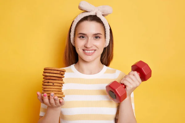 Delighted woman with hair band in striped T-shirt holding red dumbbell and sweet cookies, suggesting to burn calories, looking at camera with toothy smile, posing isolated on yellow background.