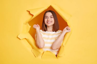 Sarı kağıt delikte çizgili tişört giymiş gülümseyen mutlu kadının yatay görüntüsü, kollarını kaldırmış, başarısını kutluyor, dişlek bir gülümsemeyle kameraya bakıyor..