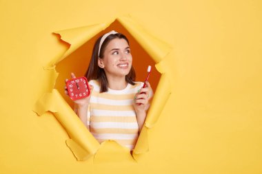 Sarı kâğıt delikte poz veren, elinde çalar saat ve diş fırçası tutan çizgili gömlek ve saç bandı takan gülümseyen bir kadının reklam ya da promosyon için fotokopi alanına baktığı yatay bir görüntü..