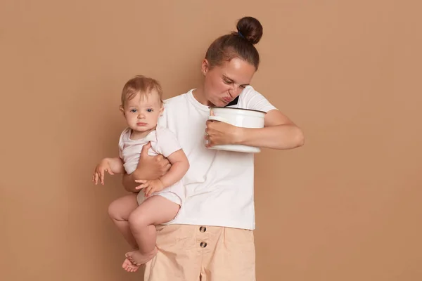 Portrait of sad woman wearing white casual style shirt holding in arms toddler daughter and and smelling spoiled food in pot while talking on cell phone isolated on brown background.