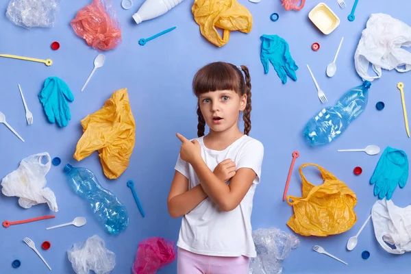 Indoor shot of shocked sad little girl wearing casual T-shirt looks sadly at camera, showing all plastic garbage, concerned by serious environmental problem, posing against blue background.