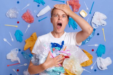 Öfkeli sinirli şaşkın kirlilikten nasıl kaçınacağını bilmiyor, insanların ekolojik olarak arkadaş canlısı olmalarını talep etmek umutsuz bir bakış, mavi duvara karşı çok fazla plastik çöp ile poz vermek, çığlık atmak