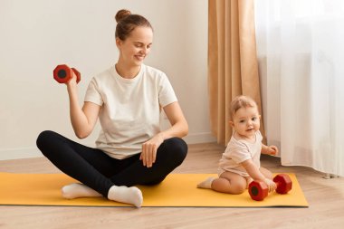 Çekici bir kadının, küçük kızıyla yoga minderinde oturup pazılar ve trisepsler için egzersiz yaparken, olumlu duygular ifade ederken, kırmızı halterle kolunu kaldırırken..