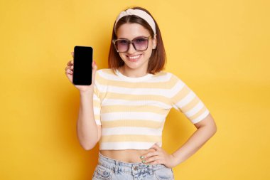 Güneş gözlüğü, çizgili tişört ve saç bandı takan, eli kalçasında duran ve cep telefonunu boş bir şekilde gösteren, sarı arka planda izole edilmiş poz veren mutlu bir kadın görüntüsü..