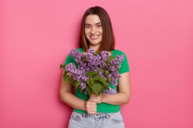 Photo of pleased dark haired woman wearing green T-shirt standing looking with pleasure at camera, carries lilac flowers, models against pink background. Spring present.
