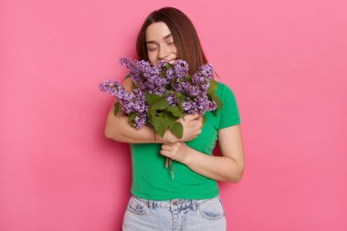 Indoor shot of satisfied woman wearing green t shirt, carries lilac flowers, embracing her bouquet,posing isolated over pink background, enjoying her present from boyfriend.