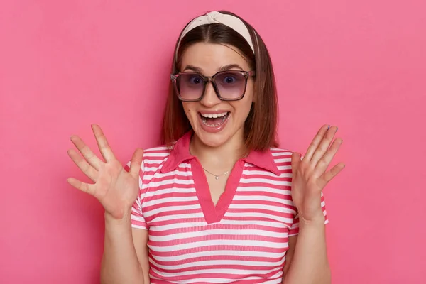 Horizontal shot of excited surprised woman wearing striped T-shirt and sunglasses posing isolated over pink background, standing with raised arms, being pleasantly surprised.
