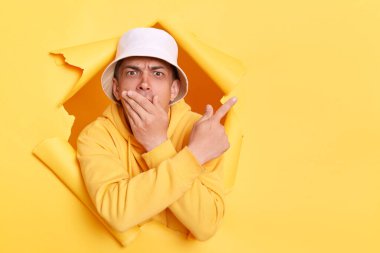 Kapüşonlu ve panamalı, ağzı palmiyeyle kaplayan, reklam alanını işaret eden, sarı kağıt duvarda yırtılan adamın yatay görüntüsü..