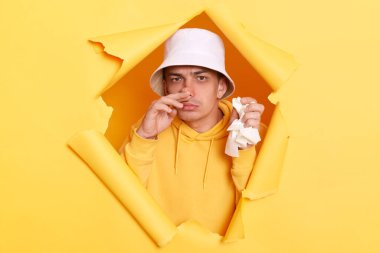 Kafkasyalı hasta, kapüşon ve panama giyen, burnu akan, bahar alerjisi olan, kameraya bakan, sarı kağıt duvardaki delikte poz veren genç bir adamın resmi..