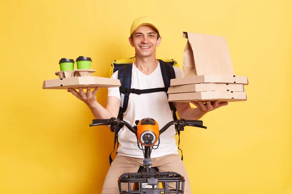 Positive courier man on bike being in hurry to delivery food to clint, holding carton pizza boxes and disposable cups with hot beverage, wearing white t shirt and cap, isolated over yellow background.