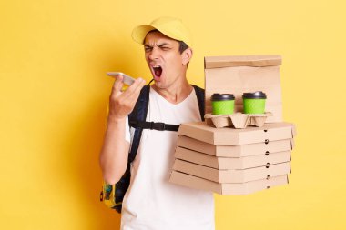 Portrait of angry man courier with thermo backpack in white T-shirt and cap, recording voice message to client, frowning face, delivering pizza and coffee, isolated over yellow background.