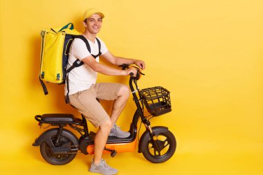 Sarı arka planda izole edilmiş beyaz tişört ve kapaklı termo sırt çantalı bir erkek kuryenin tam boy profil portresi. Elektrikli bisiklete biniyor, müşterilere siparişleri teslim ediyor..