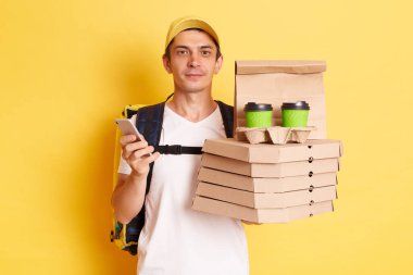 Horizontal shot of serious calm man courier with thermo backpack in white T-shirt and cap, looking at camera, holding client's order, standing with smart phone in hands isolated over yellow background