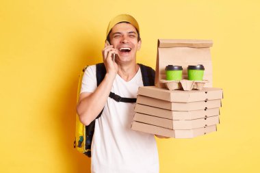 Beyaz tişörtlü ve şapkalı mutlu gülen adam kuryesinin kapalı çekimleri sarı arka planda izole edilmiş telefon konuşmaları pizza kutuları ve kahve paketleri..