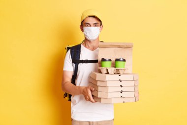 Indoor shot of man courier with thermo backpack in white T-shirt, cap and protective mask isolated over yellow background, holding delivering order, working during flu epidemic.