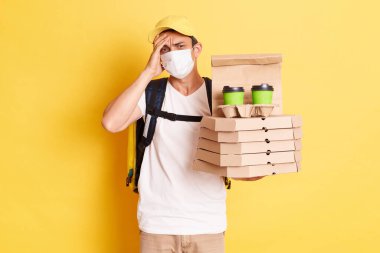 Indoor shot of tired deliveryman in protective mask standing against yellow wall, holding pizza and coffee, suffering headache, forgot clint address, working during quarantine.
