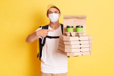 Portrait of deliveryman with thermo backpack in white T-shirt, cap and protective mask standing against yellow wall, pointing at pizza boxes and disposable cups with coffee.