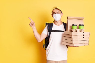 Image of positive deliveryman with thermo backpack in white T-shirt, cap and protective mask standing against yellow wall, holding food order, pointing at copy space for advertisement.