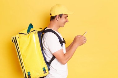 Side view portrait of positive handsome deliveryman with thermo backpack in white T-shirt and cap standing against yellow wall, standing using cell phone, chatting with client.