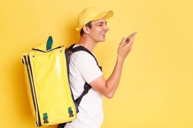 Profile portrait of smiling happy deliveryman with thermo backpack in white T-shirt and cap using smart phone for recording voice message, standing against yellow wall.