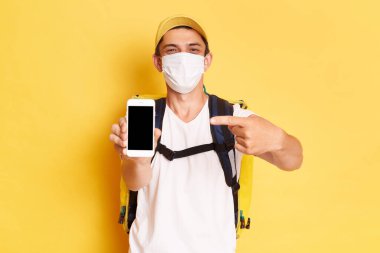 Beyaz tişört ve kapaklı termo sırt çantalı teslimatçının yatay görüntüsü sarı duvara yaslanmış cep telefonuyla reklam veya tanıtım metni dağıtırken boş ekranı gösteriyor..