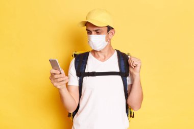 Beyaz tişörtlü ve kapaklı termo sırt çantalı bir teslimatçının sarı duvara dayanmış cep telefonu kullanırken, ekrana bakarken, karantina, grip ya da covid salgını sırasında çalışırken çekilmiş bir fotoğrafı..