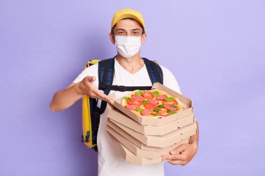 Portrait of courier man wearing white T-shirt and protective with thermo backpack holding pizza boxes, delivering food during quarantine, posing isolated over purple background.