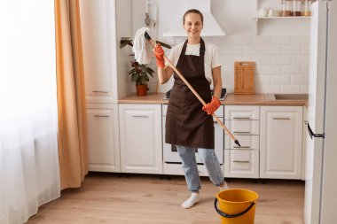 Portrait of adorable hardworking female wearing brown apron and rubber gloves making fast domestic work in white kitchen, holding mop, looking at camera with positive expression.