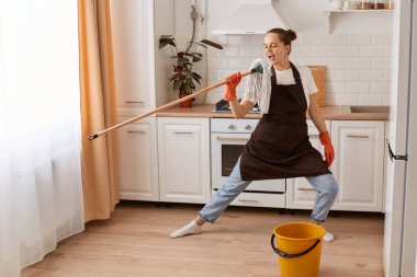 Indoor shot of happy positive housewife cleaning her flat, washing floor, having fun while doing household chores, wearing casual clothing, gloves and brown apron.