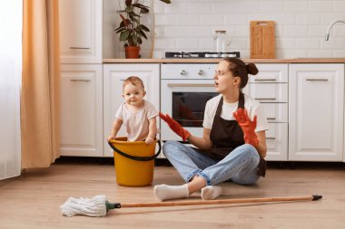 Küçük kızıyla ev işi yapan korkmuş bir kadının uzun bir portresi mutfakta yerleri yıkıyor, kovadaki çocuğa bakıyor, ellerini açıyor..