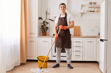 Indoor shot of smiling positive female wearing brown apron and rubber gloves, holding mop, washing floor in kitchen, looking at camera, being in good mood while doing household chores.