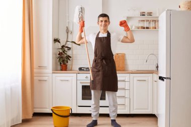 Portrait of handsome man wearing brown apron and orange gloves, finishing his cleaning the house, washing floor in kitchen, holding mop, raised arms, showing his power.