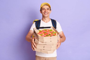 Çekici, beyaz bir teslimatçı tişört giyiyor ve elinde pizza kutuları, müşteriye düzen getiriyor, kameraya gülümsüyor, mor arka planda izole bir şekilde poz veriyor..