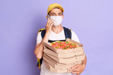 Courier man wearing t shirt and protective mask holding pizza boxes in hands, posing isolated over purple background, talking on mobile phone, specify the delivery address of the order.