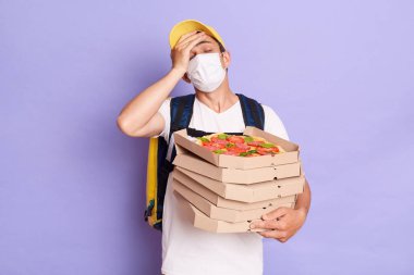 Young delivery man wearing t shirt and protective mask holding pizza boxes in hands, forgot client's address, showing facelpalm gesture, posing isolated over purple background.