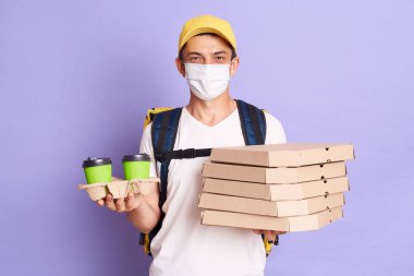 Delivery man wearing t shirt and cap holding pizza boxes and takeaway coffee in hands, delivering food during flu epidemic, bringing orders in time, posing isolated over purple background.