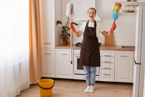 Portrait of excited woman wearing brown apron making fast domestic work wiping and washing floor in modern kitchen, being in good mood, raised arms with her equipment, finishing her cleaning.
