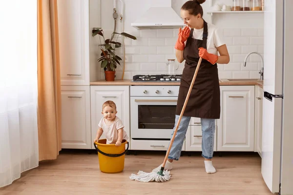 Full length shot of nice attractive hardworking woman making fast domestic work with her cute baby daughter in bucket, wiping floor in modern light white interior kitchen.