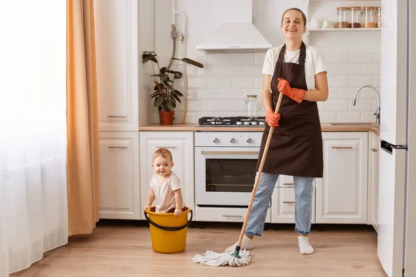 Full length shot of cheerful cheery housewife making fast domestic work wiping floor in kitchen, spending time with her baby while washing,m ho;ding mop, wearing brown apron, looking at camera.