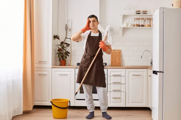 Full length portrait of tired man in rubber gloves and brown apron having a rest from cleaning kitchen floor, standing with exhausted facial expression, wiping his forehead, doing household chores.