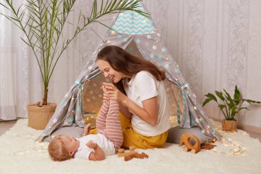 Kapalı alanda gülümseyen mutlu anne pietee çadırında oynarken yerde oturup bebek bebekle eğlenirken, anne kucağında çocuk ayaklarıyla.
