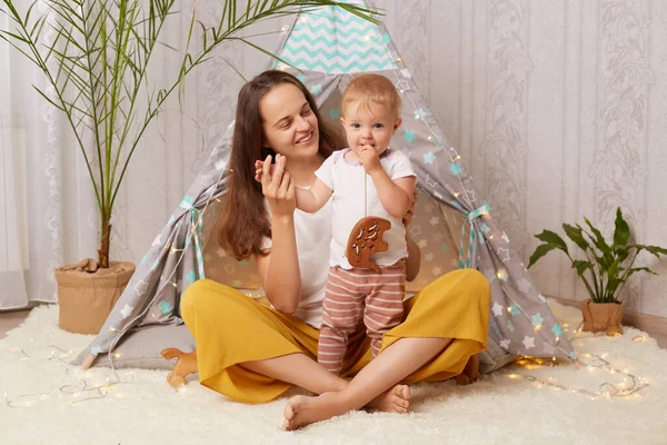 Indoor shot of smiling positive woman with her kid, baby girl and her mother playing at home in peetee tent while sitting on the floor, family spending time together, female enjoying maternity leave.