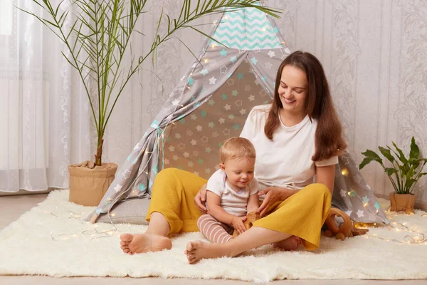 Portrait of mother playing at home in wigwam with her baby daughter while sitting on the floor, female looking at toddler kid with love and gentle, happy motherhood and childhood.