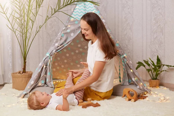 Indoor shot of young mother playing with with baby daughter on floor near kids wigwam, expressing positive emotions, spending time with cute kid in peetee tent at home.