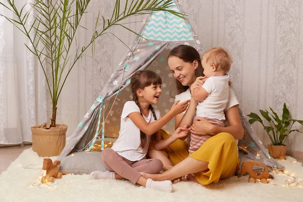 Indoor shot of Caucasian attractive woman with her children sitting together teepee tent, mom playing with daughters, spending weekend with kids while playing in wigwam.