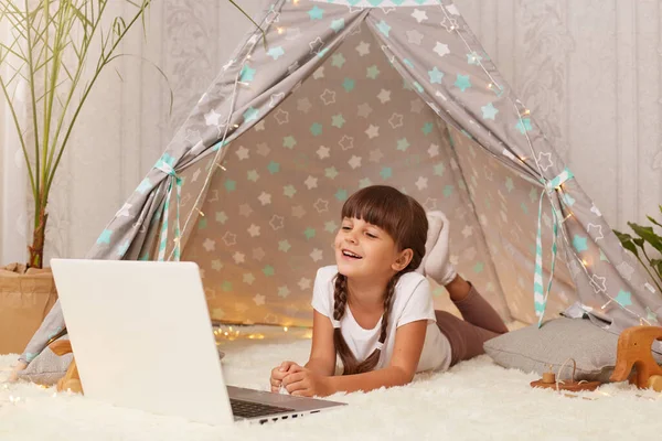 Indoor shtot of dark haired little girl with pigtails lying in peetee and watching having online conversation on laptop, looking at device display with satisfied facial expression.