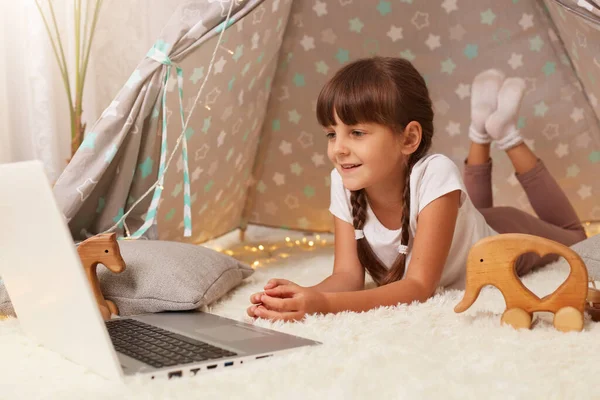 Horizontal shot of cute delighted little girl with braids wearing casual clothing laying on floor on soft carpet in wigwam and watching cartoon on laptop, spending free time alone at home.